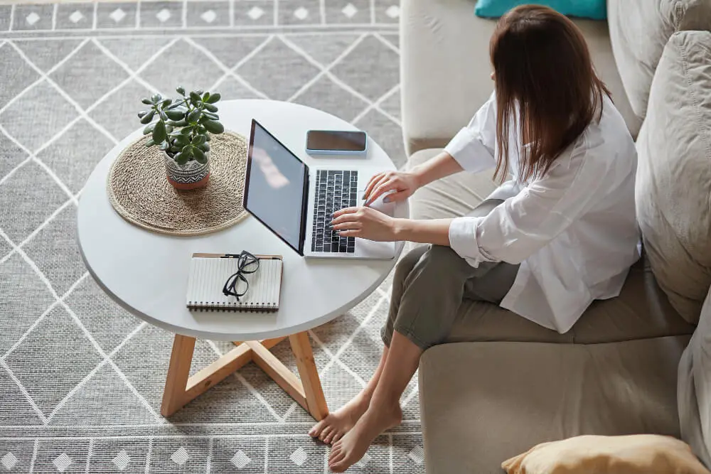 Kobieta siedząca na kanapie przy stoliku kawowym. Na stoliku otwarty laptop, na którym ona pracuje. Na podłodze szary dywan.