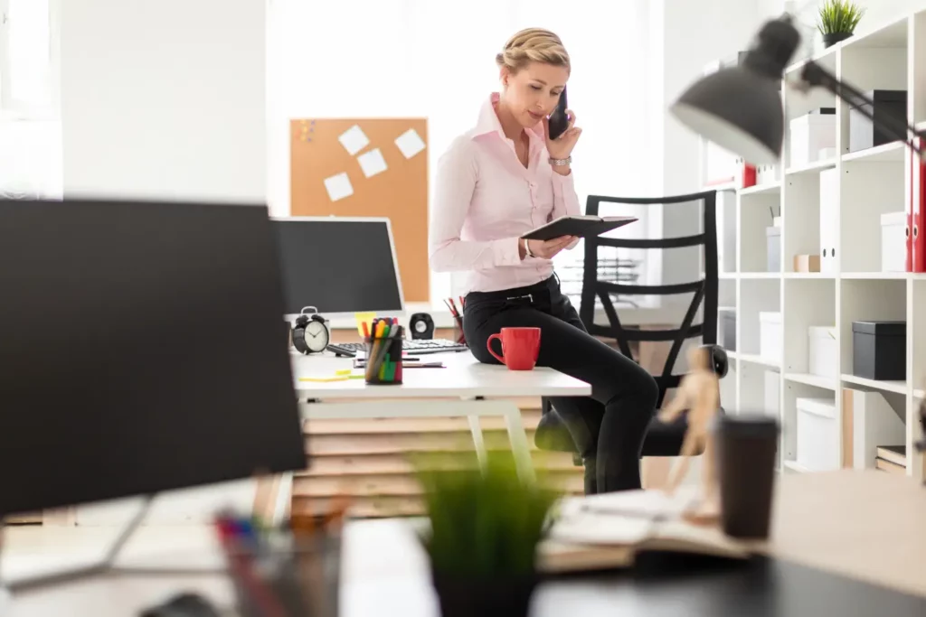 Kobieta w eleganckiej koszuli i spódnicy w biurze, siedzi na blacie biurka i spogląda w notes rozmawiając przez telefon.